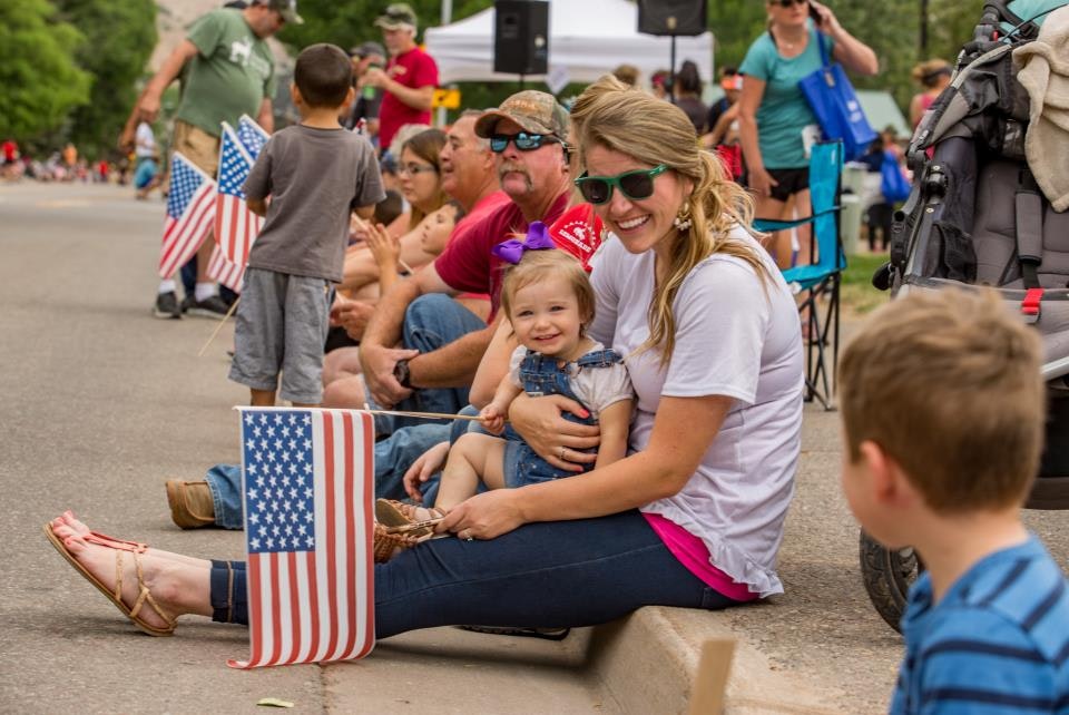 People sitting on a curb with American flags, including a woman holding a smiling child.