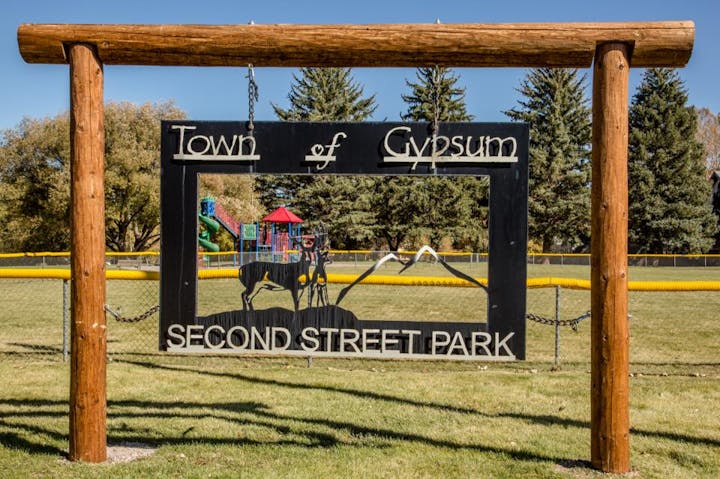 Sign for "Town of Gypsum, Second Street Park" with playground and trees in the background.