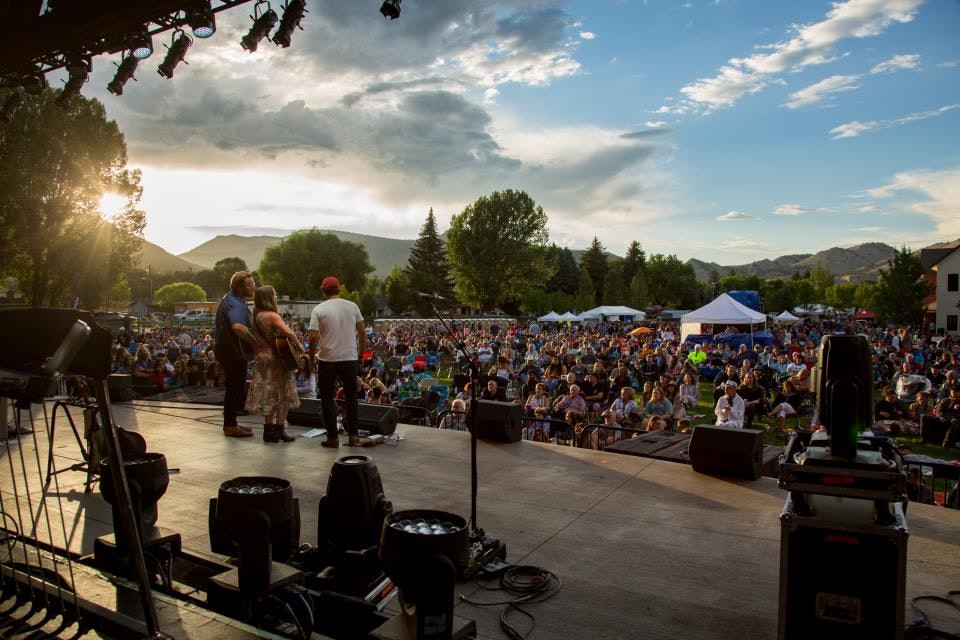 Performers on stage with a large outdoor audience and scenic mountain backdrop during sunset.