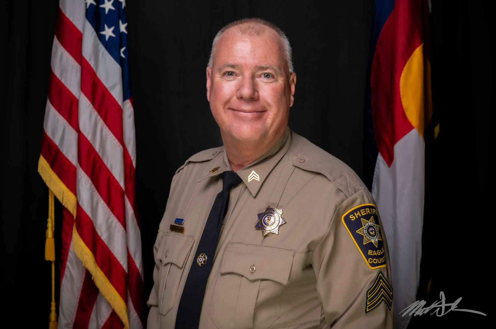 A uniformed sheriff poses in front of two flags, smiling, with sheriff’s badge visible.