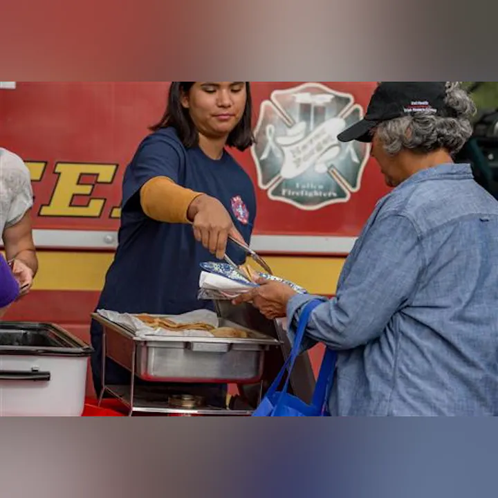 A woman at a food service station hands food to a customer, with a fire department backdrop.