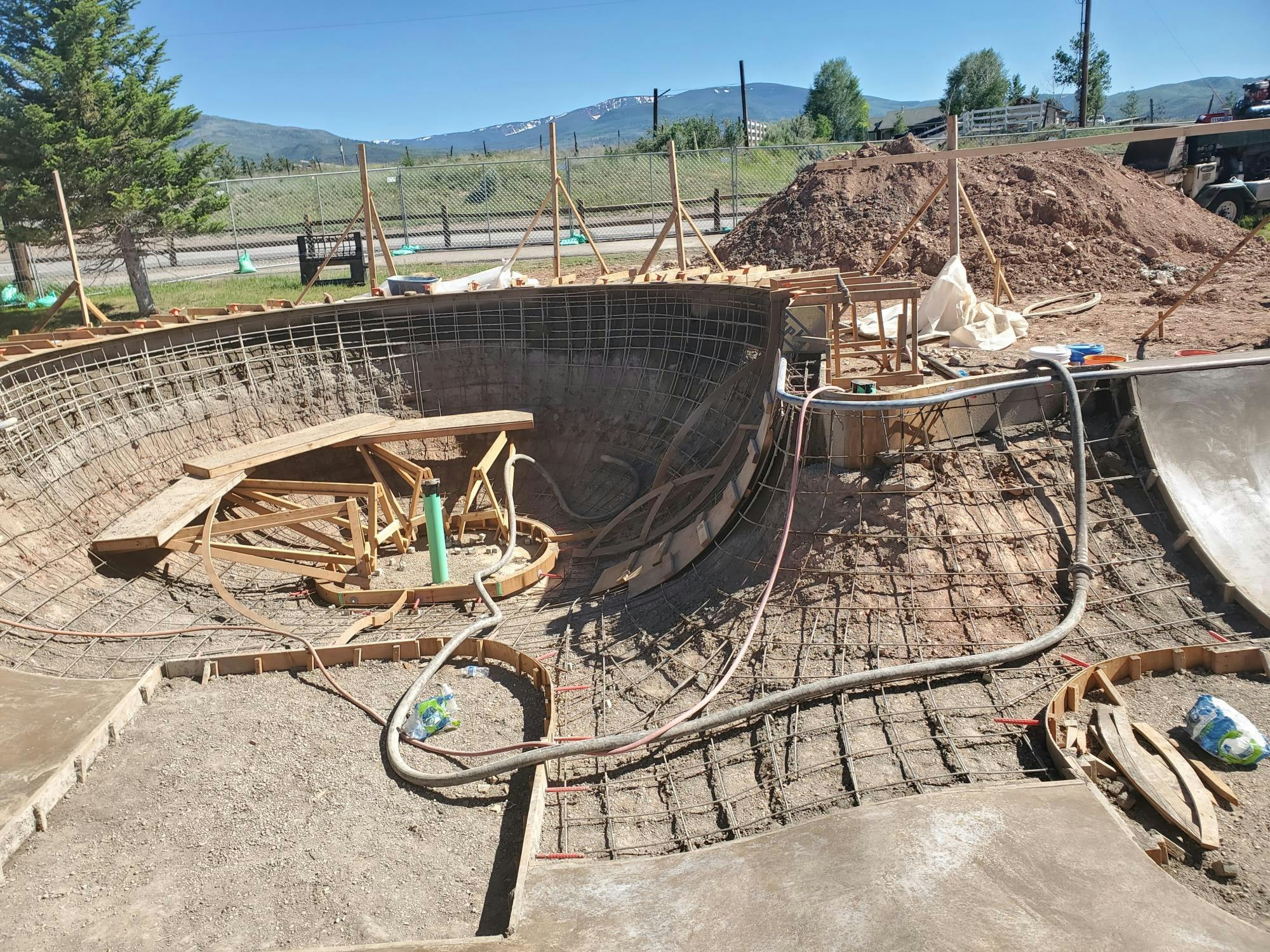 Construction site for a skatepark bowl with rebar and wooden framework, dirt piles, and mountains in the background.