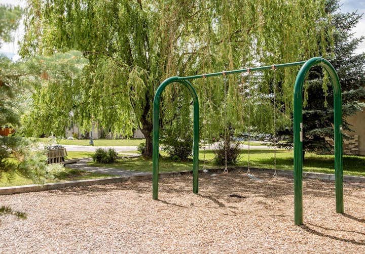 A playground with green swings, wood chips ground, surrounded by lush trees and grass. There's a bench and a path in the background.