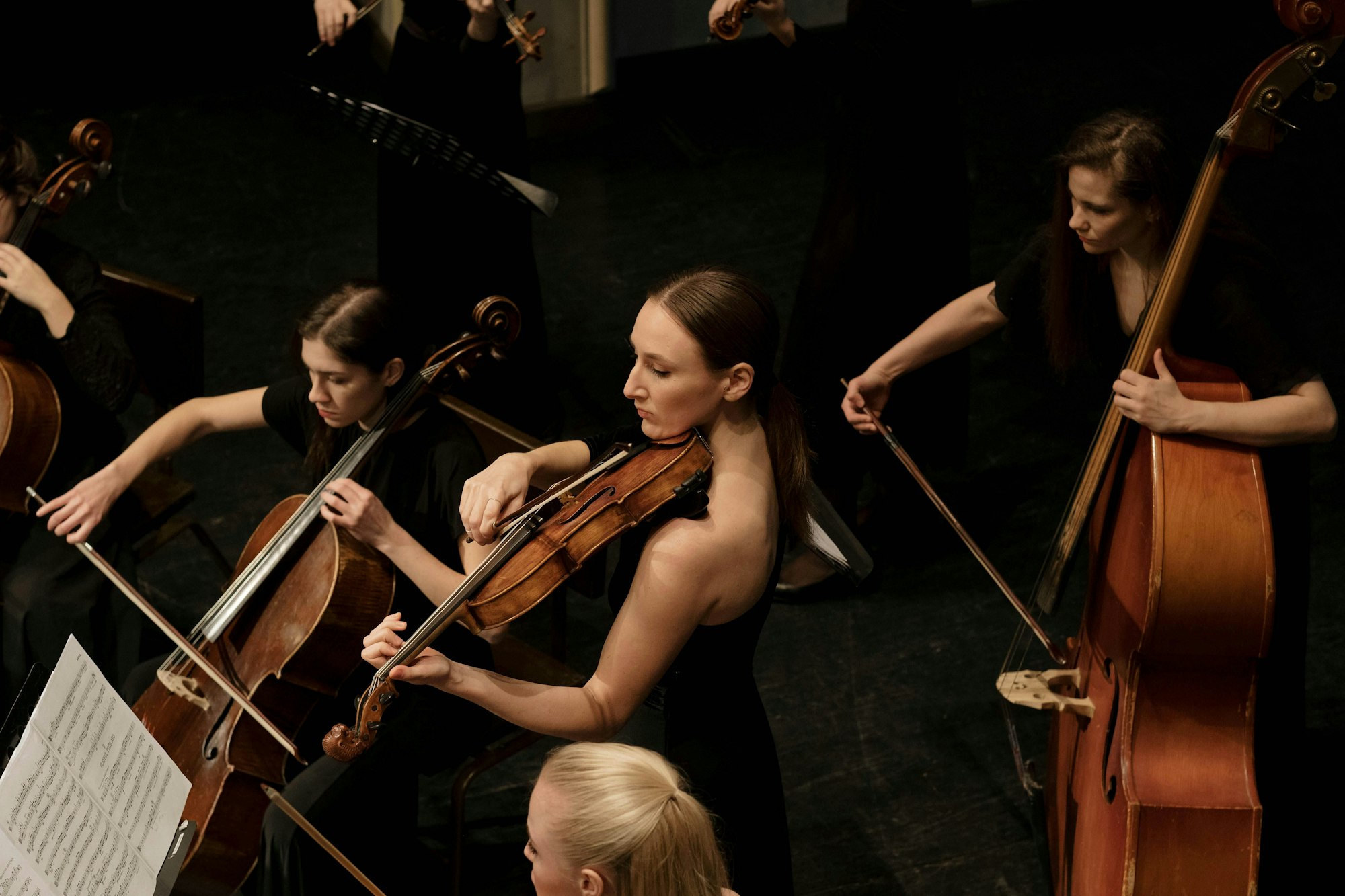 A string orchestra performs on stage, featuring musicians playing violins, violas, and cellos, all in formal attire.