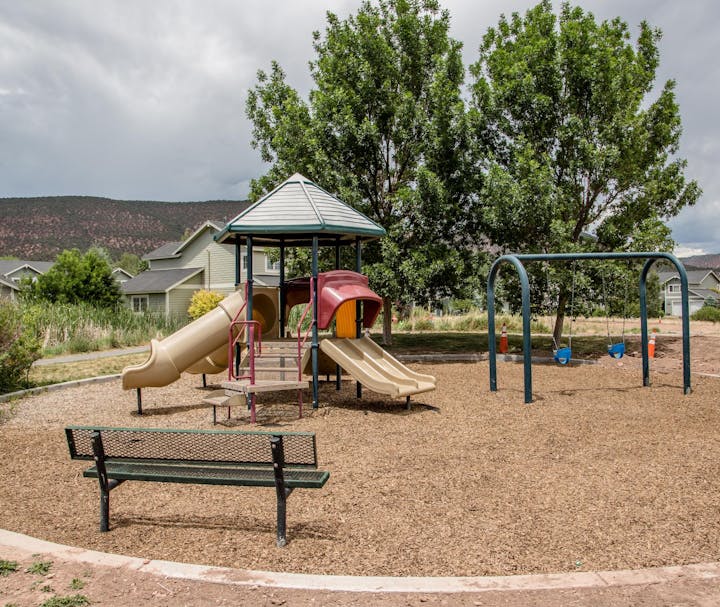 A playground with slides, swings, and a bench, surrounded by trees and houses under a cloudy sky.