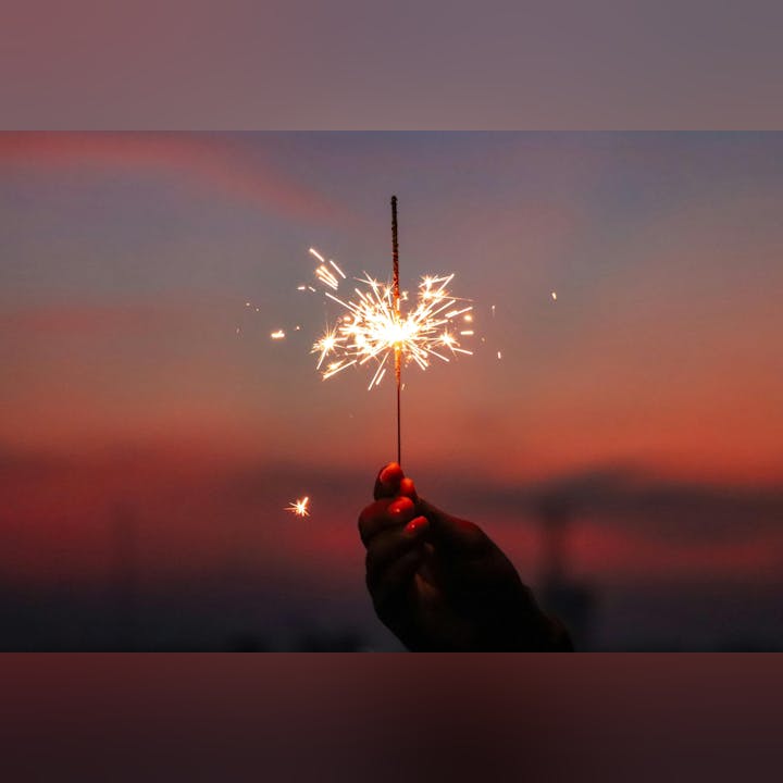 A hand holds a sparkler, emitting bright sparks against a colorful sunset background.