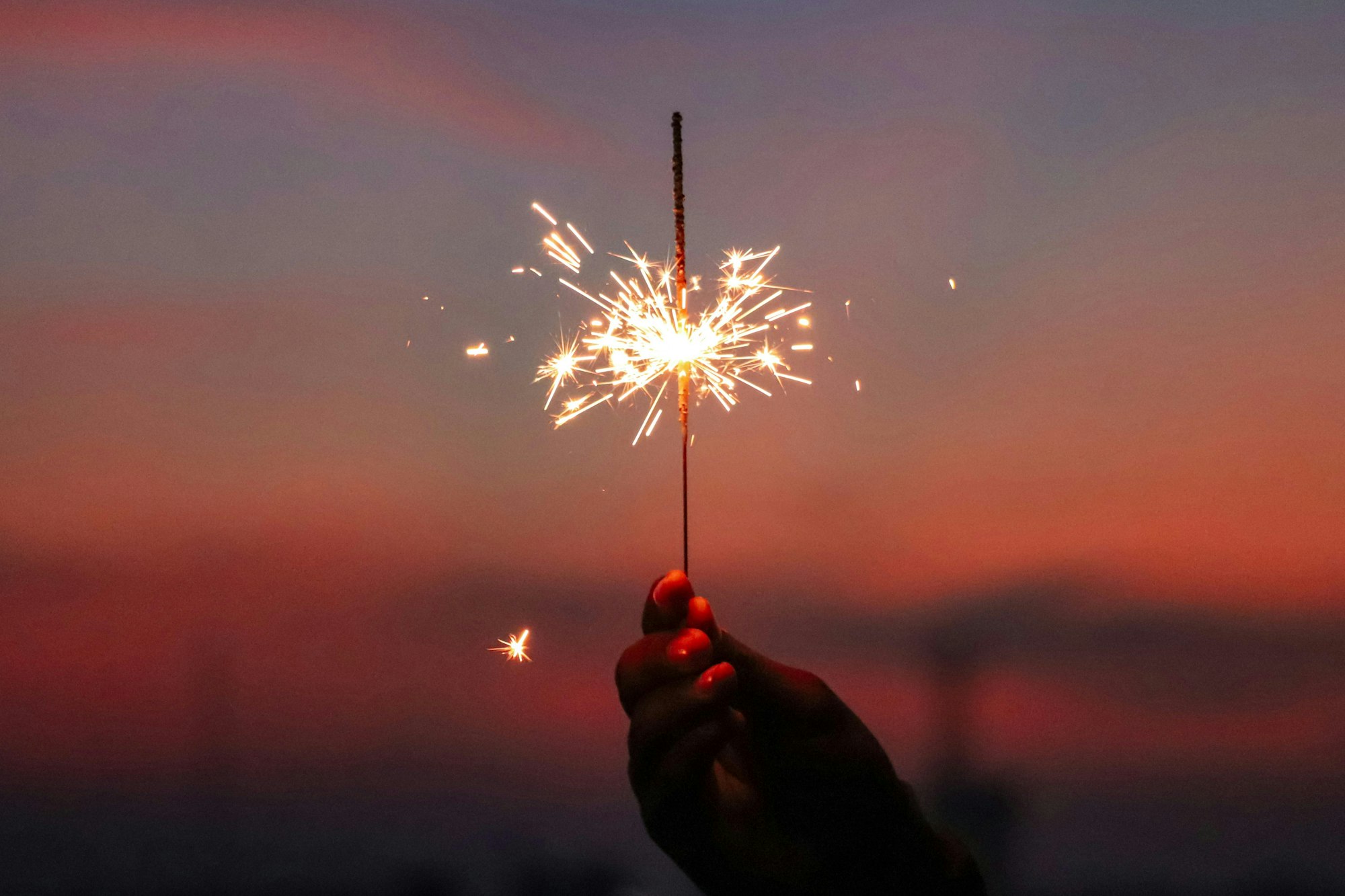 A hand holds a sparkler, emitting bright sparks against a colorful sunset background.