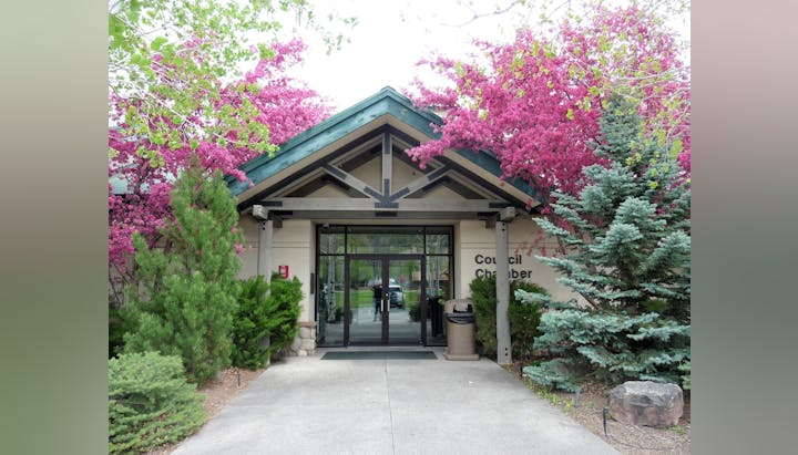 Entrance to a Council Chamber with a gabled roof, surrounded by pink blossoming trees and green shrubs.