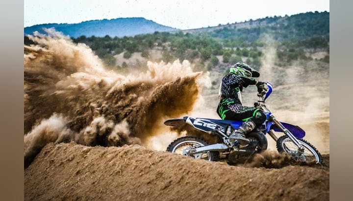 A motocross rider on a blue dirt bike kicking up a large cloud of dirt while racing on a sandy track.