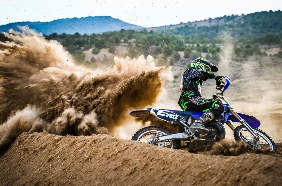 A motocross rider on a blue dirt bike kicking up a large cloud of dirt while racing on a sandy track.