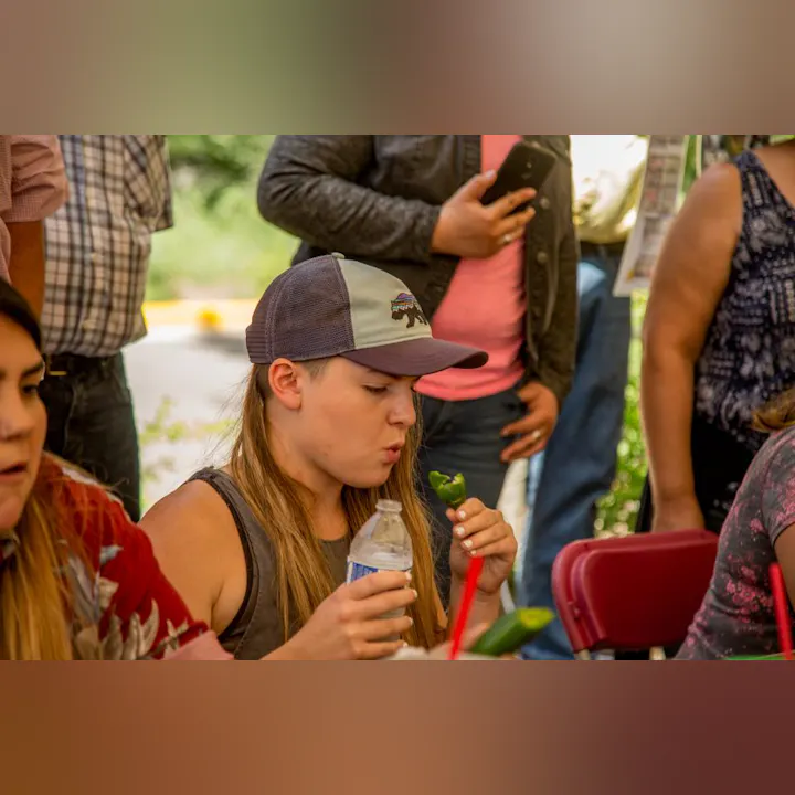 A person sitting and holding a jalapeño and a water bottle, surrounded by people.