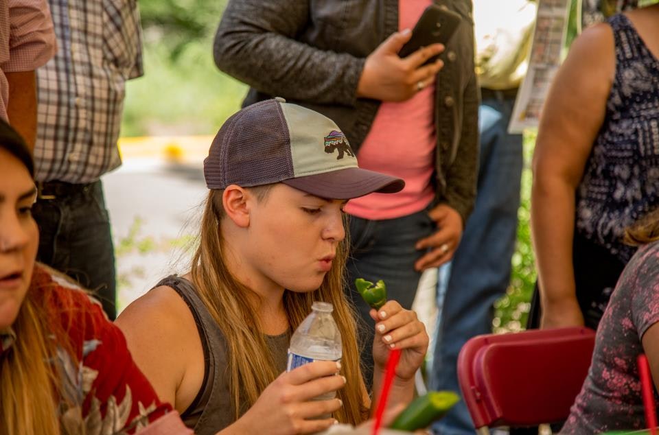 A person sitting and holding a jalapeño and a water bottle, surrounded by people.