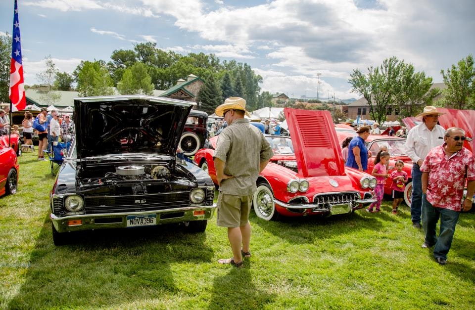 Car show with vintage vehicles, people viewing cars, open hoods, green grass, and an American flag.
