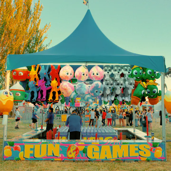 A colorful game booth at a fair, featuring stuffed animals, prizes hanging from a tent, and people enjoying the festivities.