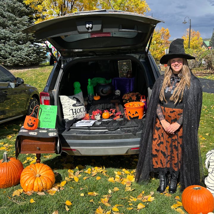 A woman in a witch costume stands by a decorated car trunk for Halloween, featuring pumpkins and spooky decorations.