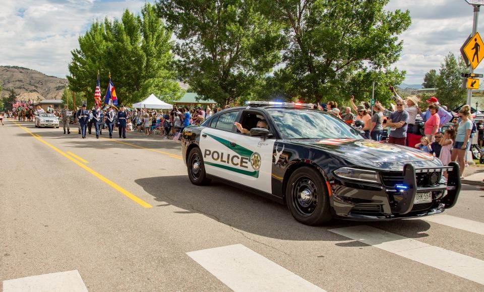Police car leading a parade with people watching, officers marching, and trees lining the street.