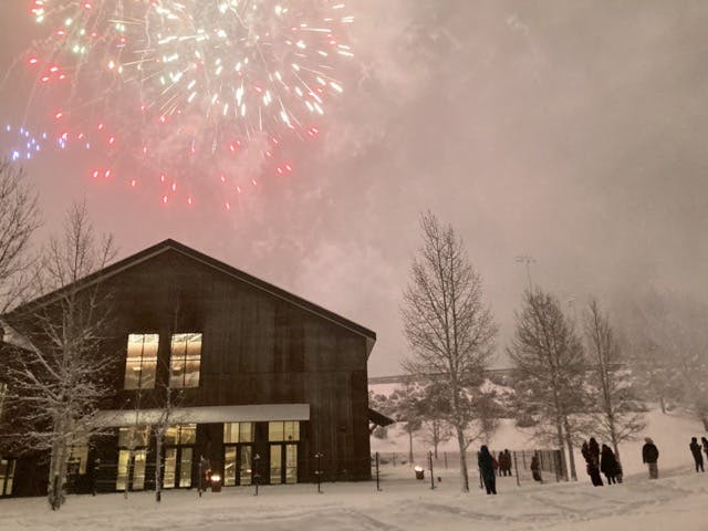 Fireworks light up the snowy sky above a wooden building and trees, with people watching from below.