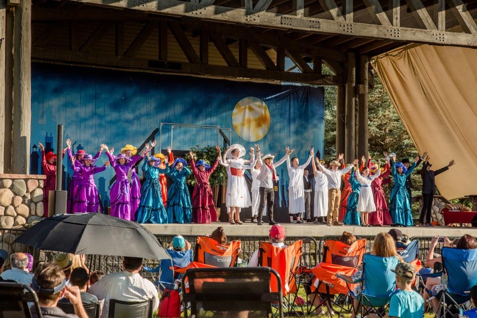 A group of people on an outdoor stage in colorful costumes, raising their hands, with an audience seated in chairs under umbrellas.
