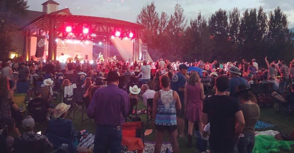 Outdoor concert with a large crowd, stage lit in red, people sitting and standing, many wearing hats, trees in the background.