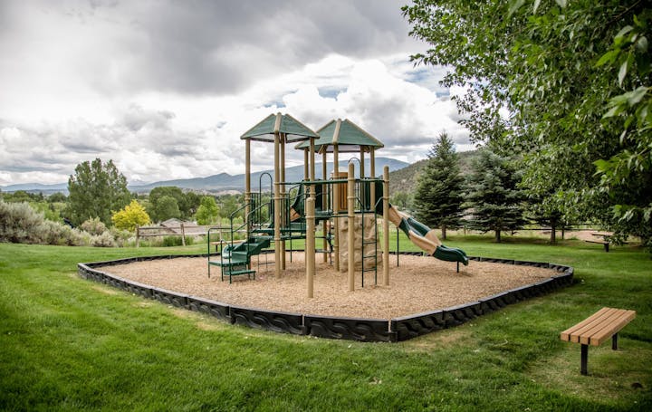 Playground with slides and climbing structures on a grassy field, surrounded by trees and mountains, with a bench nearby.