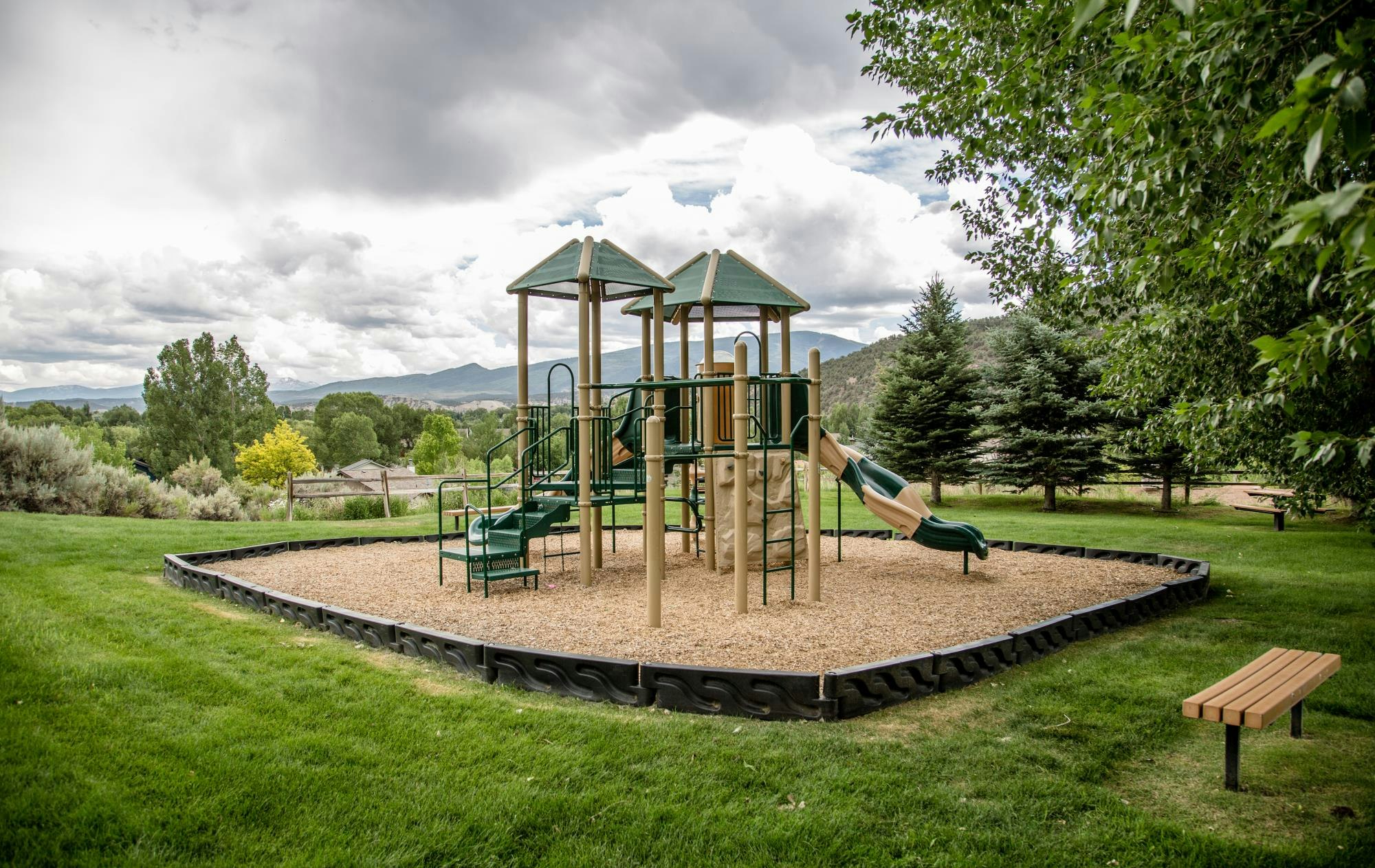 Playground with slides and climbing structures on a grassy field, surrounded by trees and mountains, with a bench nearby.