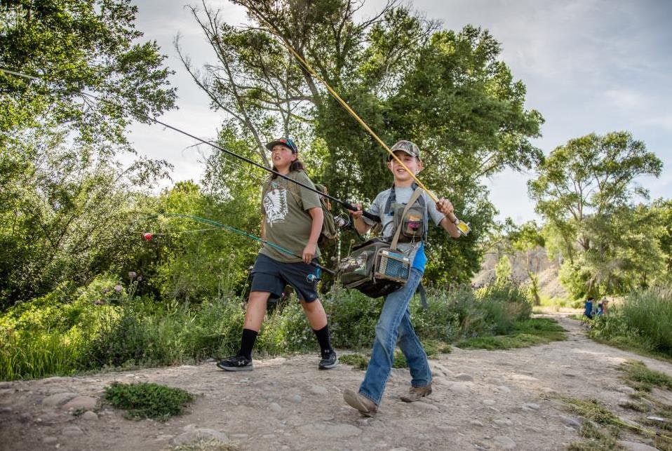 Two people walking on a path with fishing rods and gear surrounded by greenery and trees.