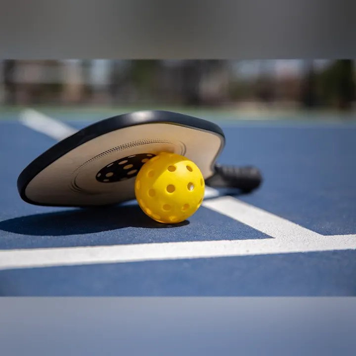 A pickleball paddle rests on a blue court next to a bright yellow perforated ball.