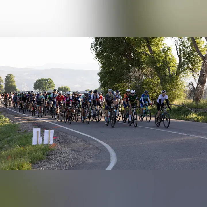 A large group of cyclists riding on a rural road, surrounded by greenery and mountains in the background, likely in a race.