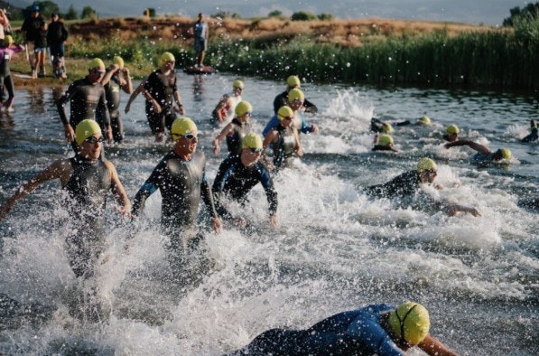A group of swimmers in wetsuits and yellow caps is starting a race in a lake, creating splashes and waves.