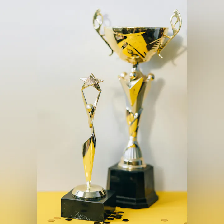 The image features two shiny trophies, one tall and one smaller with a star, set on a yellow surface with scattered coins.