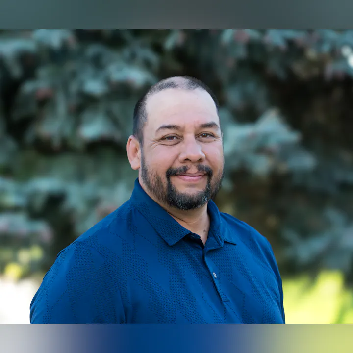 The image shows a smiling man wearing a blue polo shirt, with trees blurred in the background.