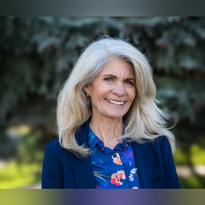 A woman with long, light gray hair smiles warmly, wearing a floral shirt and blazer, against a blurred outdoor background.