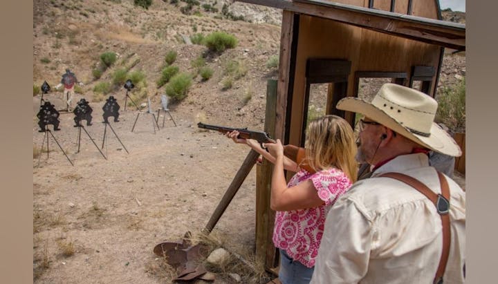 Two people at a shooting range; one is aiming a rifle at various metal targets.