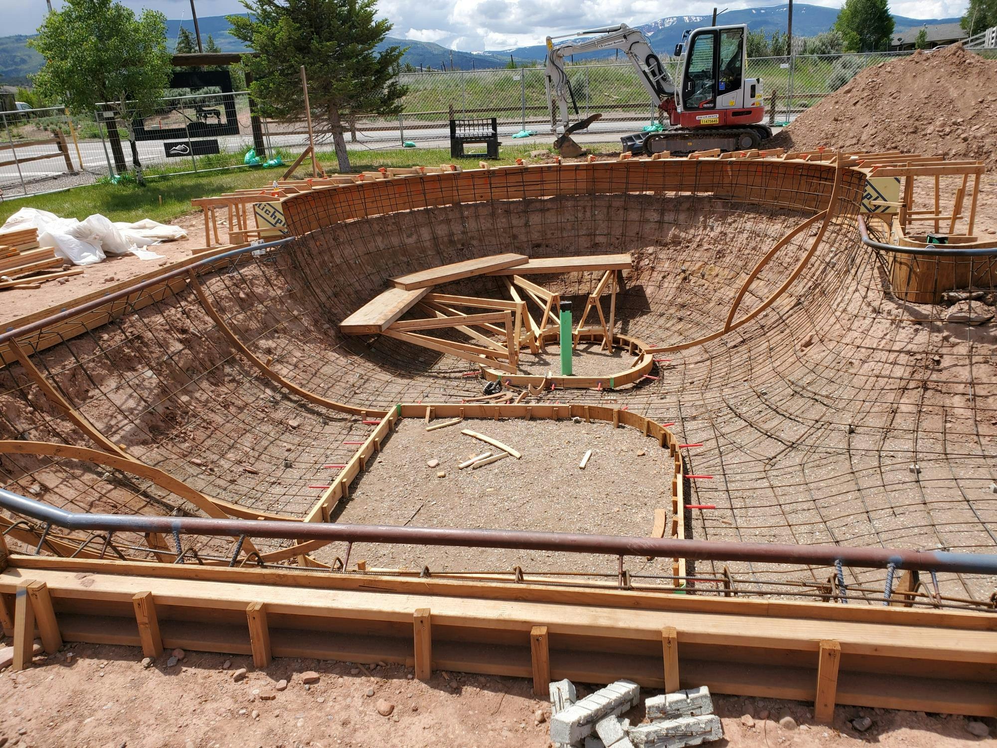 Skatepark bowl construction in progress with wooden and metal framework; an excavator and materials are nearby.