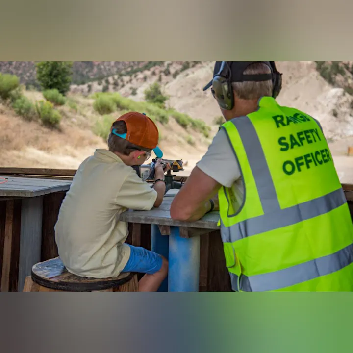 A child aiming a rifle with a Range Safety Officer supervising at an outdoor shooting range.