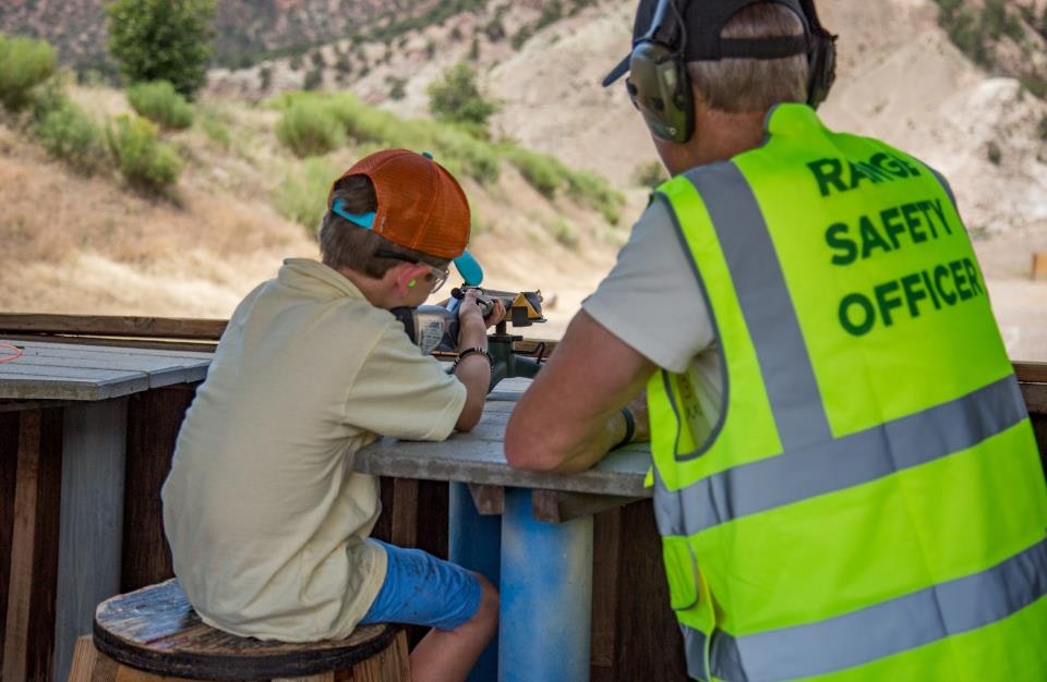 A child aiming a rifle with a Range Safety Officer supervising at an outdoor shooting range.