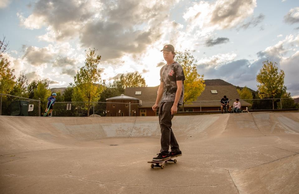 A person with a cap skateboards in a park under a cloudy sky, with others nearby, including a kid on a scooter.