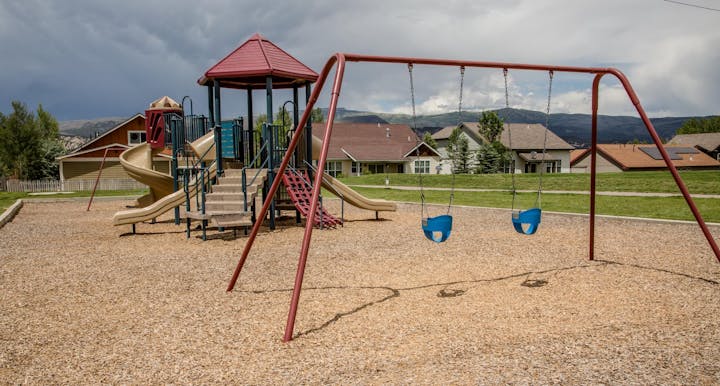 Playground with slides, swings, and climbing structures on woodchip surface. Houses and mountains in the background.