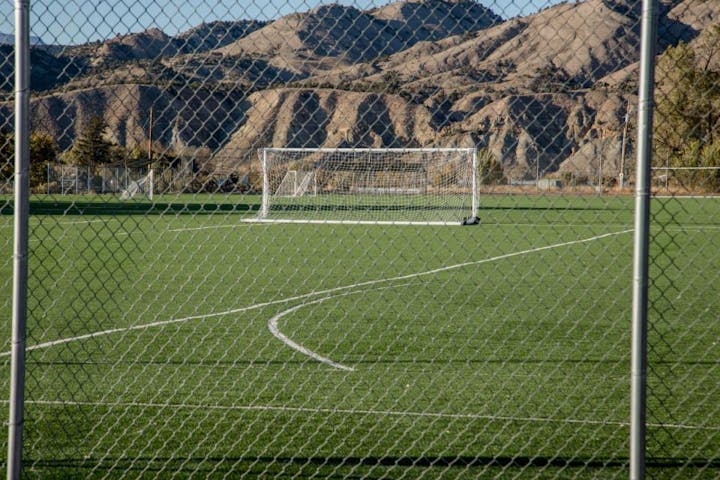A soccer field with a goal, surrounded by a chain-link fence, and mountains in the background.