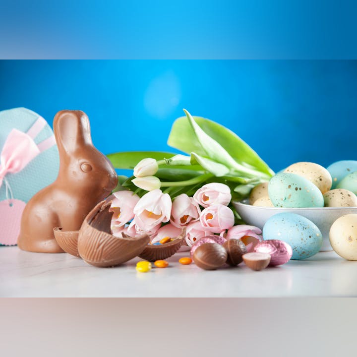 A festive Easter display featuring a chocolate bunny, colorful eggs, chocolate treats, and pink tulips against a blue background.