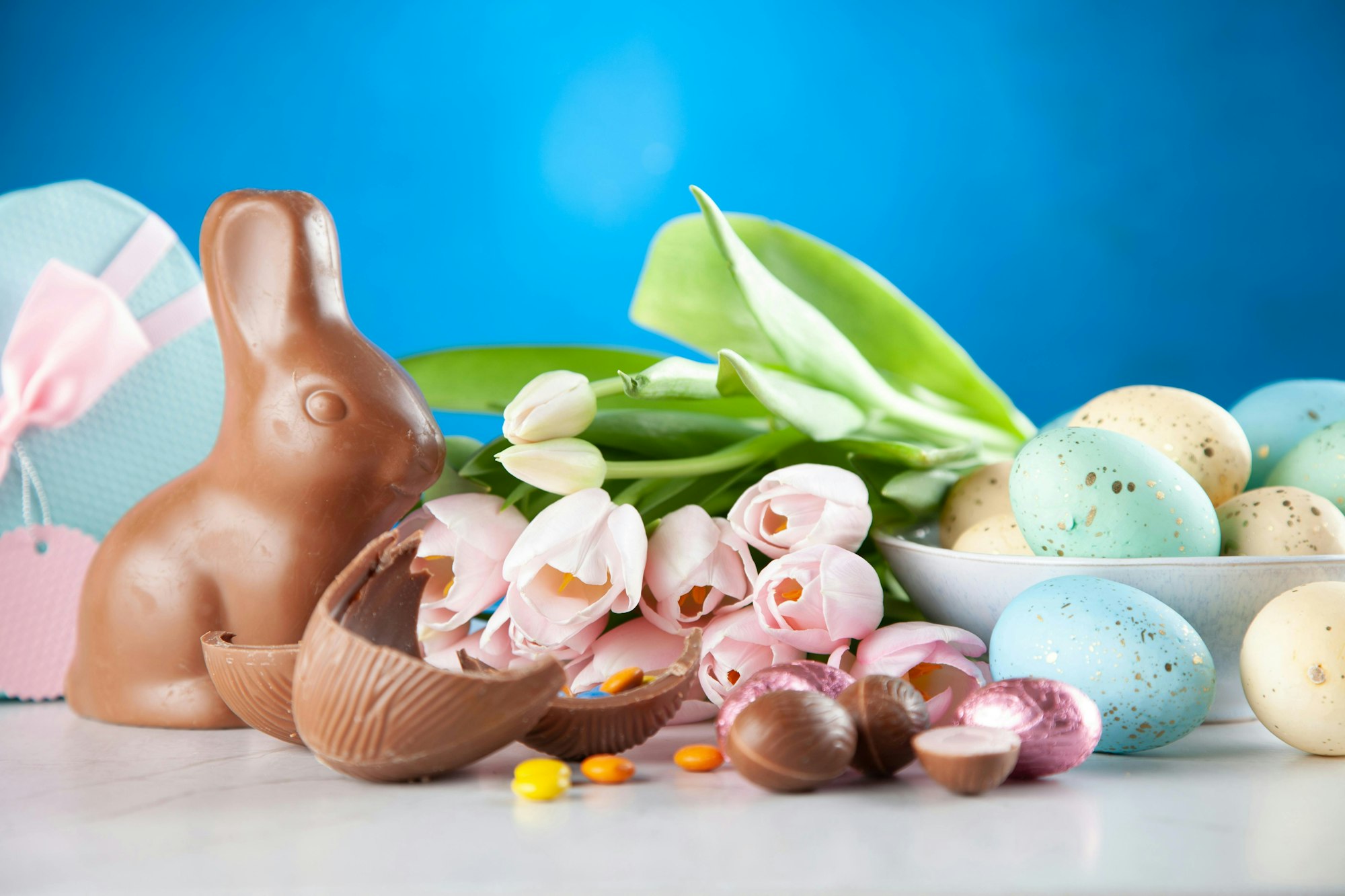 A festive Easter display featuring a chocolate bunny, colorful eggs, chocolate treats, and pink tulips against a blue background.
