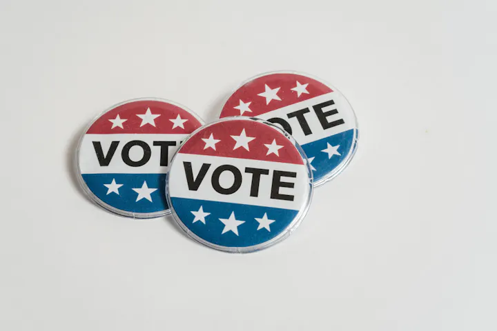 The image shows three round pins that say "VOTE," featuring red, white, and blue colors with stars.