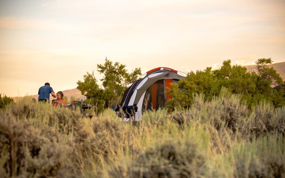 A couple is camping in a grassy area with a tent set up, surrounded by shrubs and a scenic landscape.