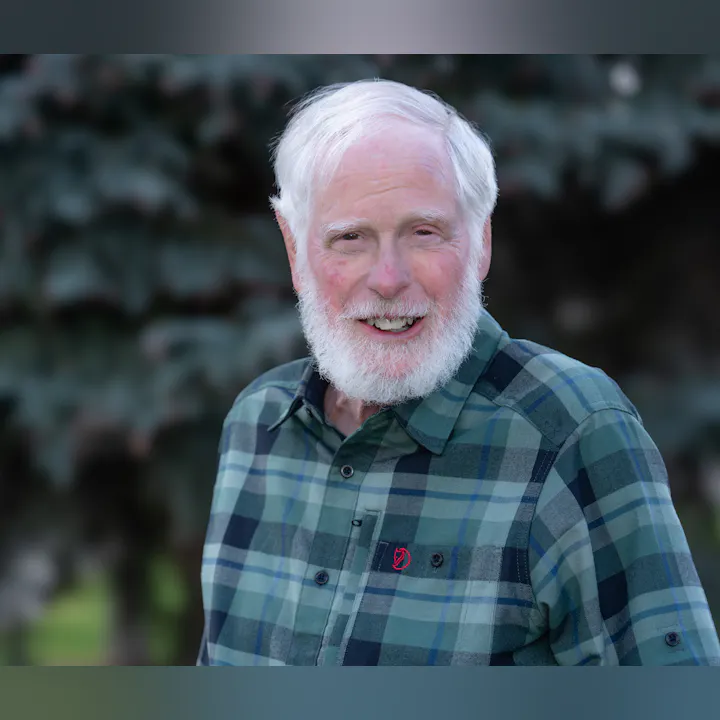 A smiling older man with a white beard is wearing a plaid shirt, standing in front of greenery.