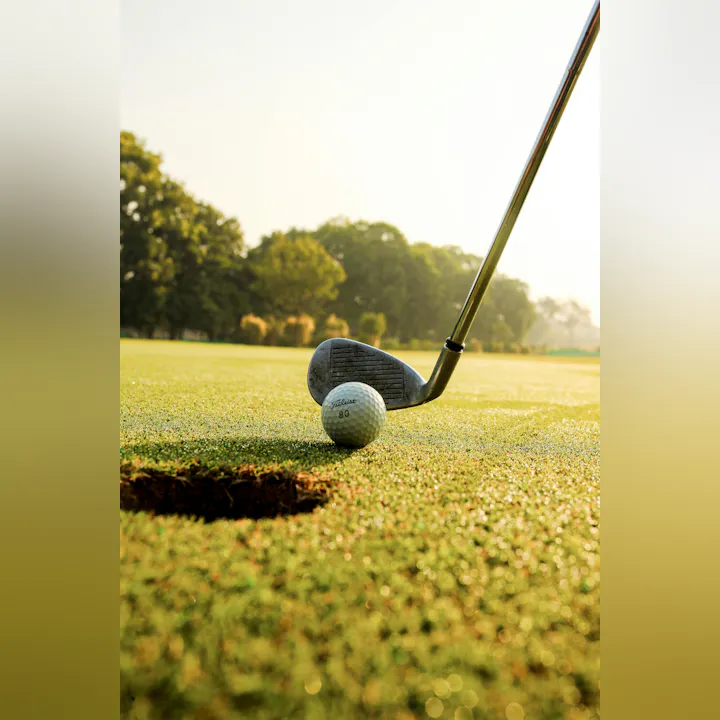 A golf club is poised to strike a golf ball near a hole on a dewy green, illuminated by sunlight.