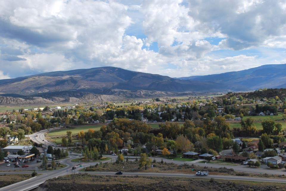 A scenic landscape with rolling hills, a small town, winding roads, and a mountainous backdrop under a partly cloudy sky.