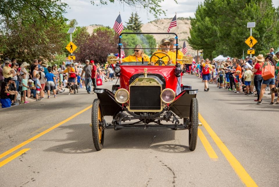 A vintage car with flags drives down a street during a crowded parade. Spectators line the road.