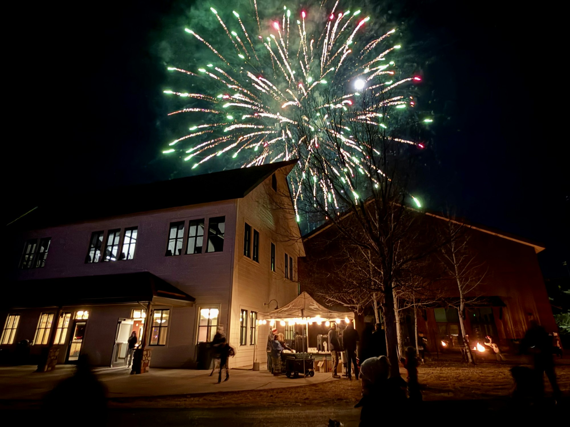 A night scene featuring colorful fireworks above buildings, with people gathered outside, enjoying the festive atmosphere.