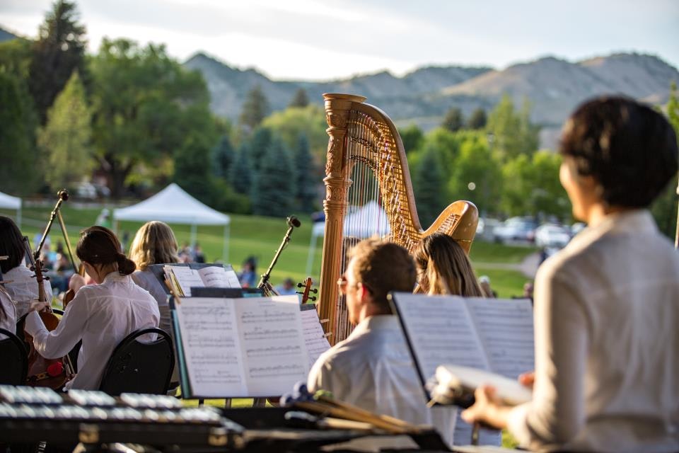 Orchestra playing outdoors with harp and musicians, sheet music visible, mountain and trees in the background.