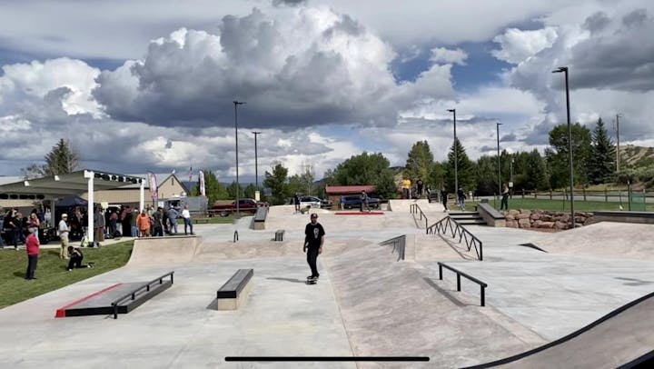 Skatepark with ramps and rails, people skateboarding. Crowd gathered under a shelter. Cloudy sky and greenery in the background.